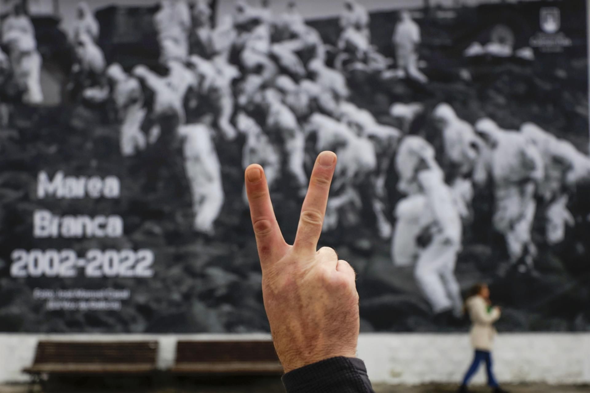 Un hombre hace la señal de victoria frente a una fotografía de voluntarios que limpiaron el chapapote del Prestigedurante el acto 'Marea Branca 2002-2022', homenaje de la Costa da Morte a los voluntarios que limpiaron las playas y rocas gallegas del chapapote que dejó el Prestige hace veinte años, evento en el que ha quedado instalada una lona de grandes dimensiones con una fotografía en recuerdo de aquellos voluntarios. EFE/Cabalar
