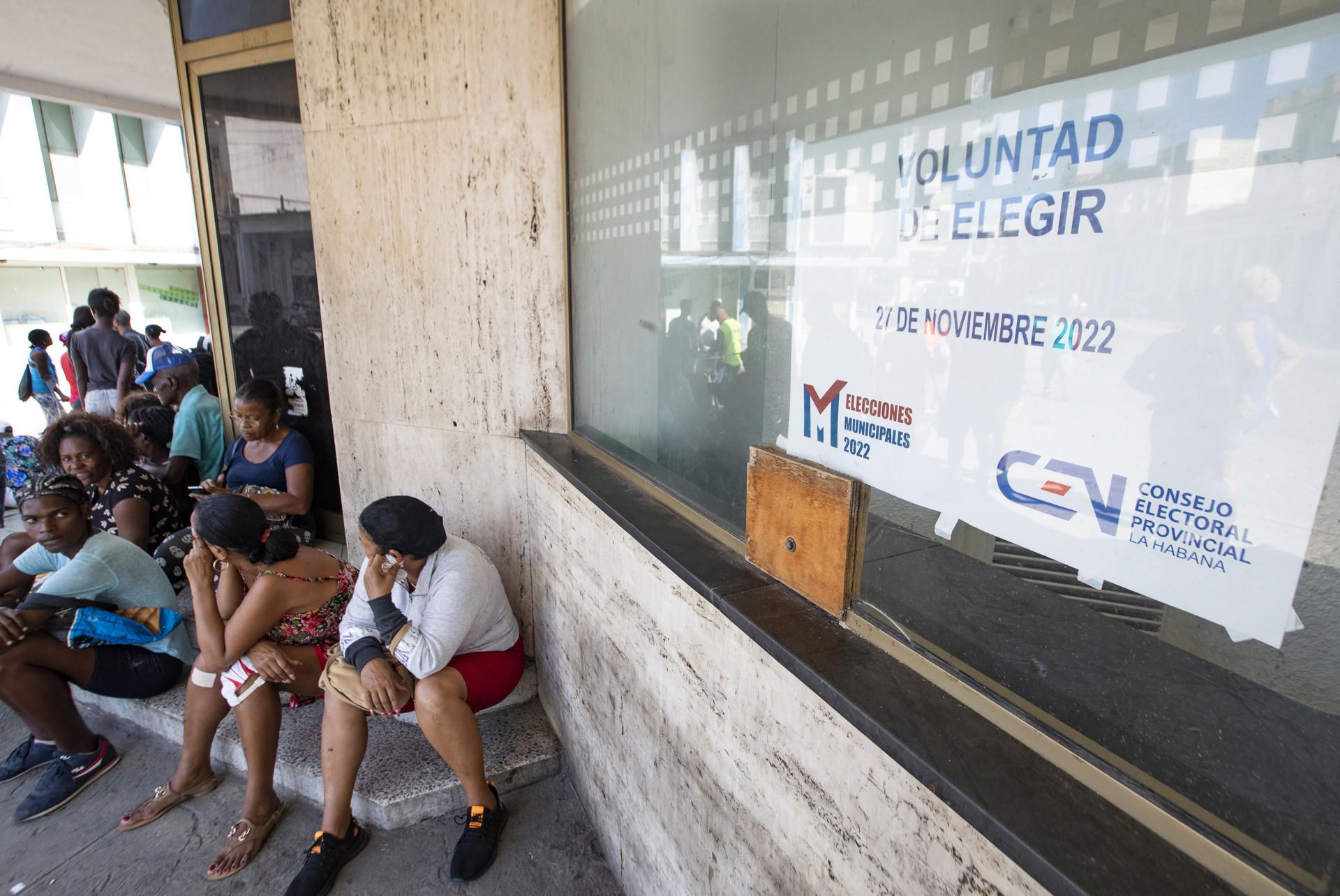 Varias personas permanecen sentadas junto a un cartel que anuncia las elecciones locales del 27 de noviembre, hoy, en La Habana (Cuba). EFE/ Yander Zamora
