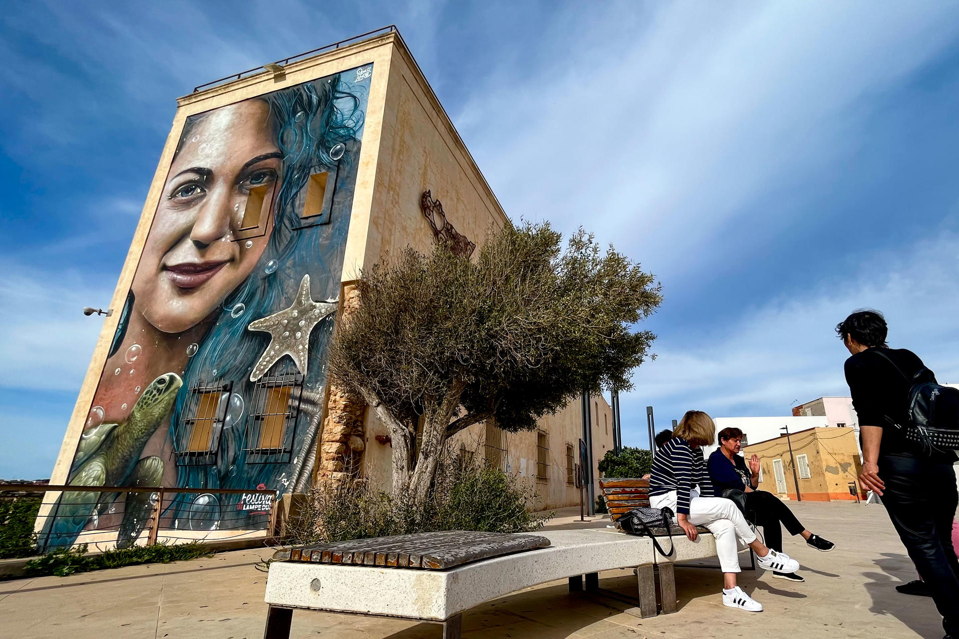 Unas mujeres charlan en la principal arteria de la isla italiana de Lampedusa, frente al puerto en el que desembarcan cada año miles de inmigrantes procedentes de la cercana África tras navegar el Mediterráneo central. EFE/Gonzalo Sánchez
