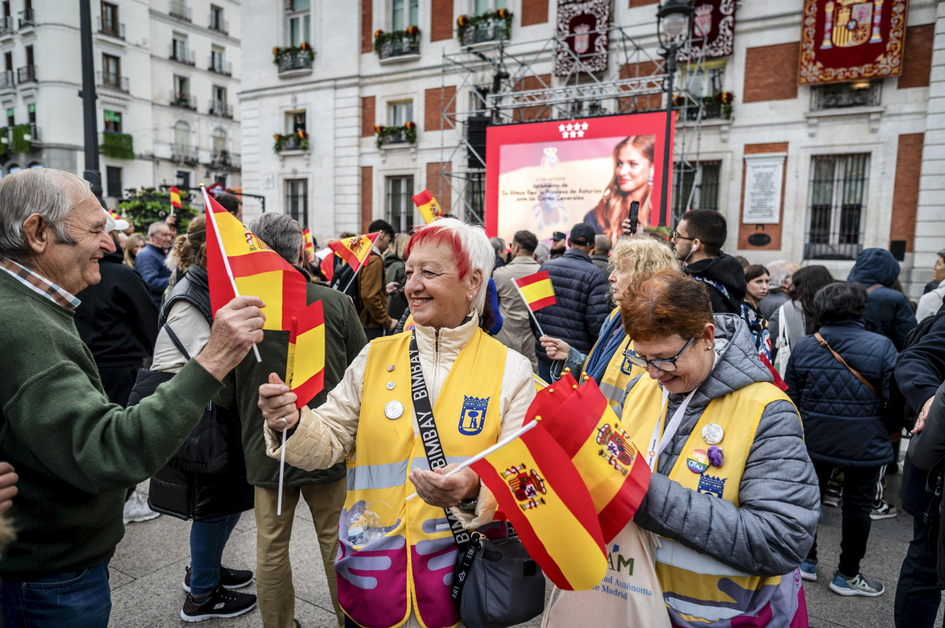 Varias mujeres reparten banderas de España frente a la Real Casa de Correos, sede del Gobierno de la Comunidad de Madrid, donde decenas de personas se congregan para la ceremonia de jura de la Constitución de Leonor de Borbón en el día de su 18 cumpleaños, un acto que representa el hito más importante de su trayectoria institucional y pavimenta el camino para que algún día se convierta en reina. EFE/ Fernando Villar
