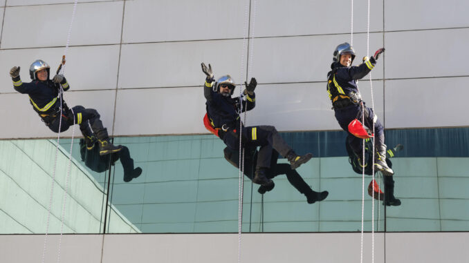 Varios bomberos saludan mientras descienden por una cuerda desde el tejado del Hospital Puerta de Hierro durante una demostración previa a la entrega de regalos a los niños ingresados en el centro médico, en Majadahonda, Madrid, este viernes. EFE/Sergio Pérez
