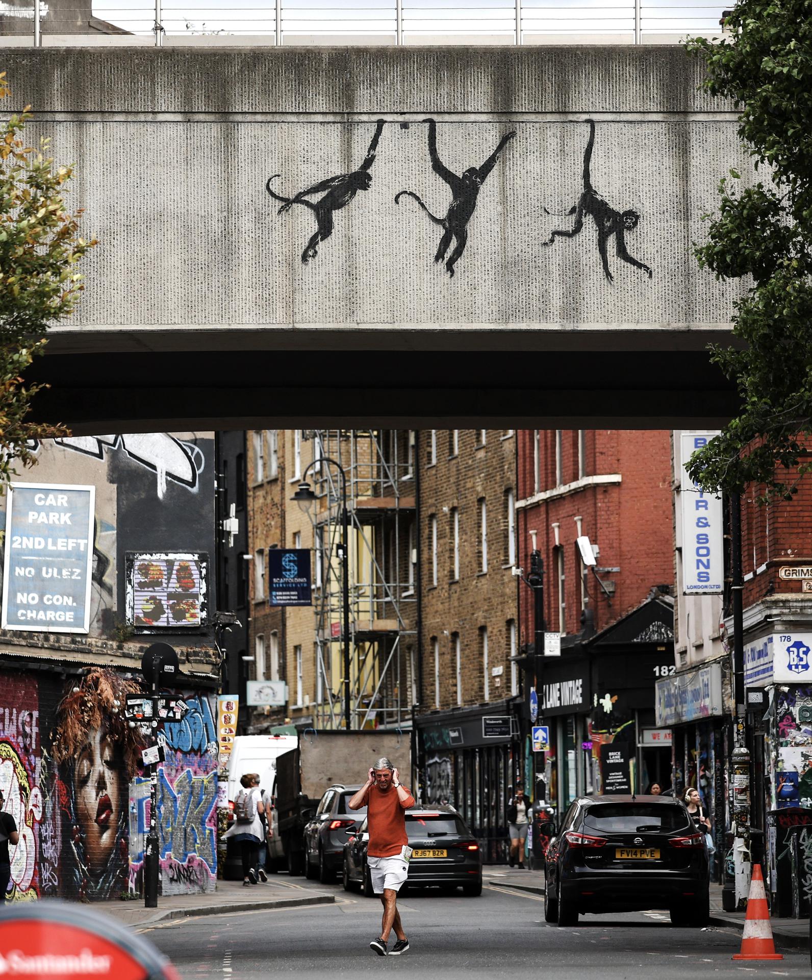 Peatones y vehículos pasan bajo un puente ferroviario Overground que muestra una nueva obra de arte de Banksy con monos en Shoreditch, al este de Londres, Gran Bretaña, el 8 de agosto de 2024. El artista británico Banksy lanzó el 7 de agosto en Londres una tercera obra de arte con temática animal en tres días. (Reino Unido, Londres) EFE/EPA/ANDY LLUVIA
