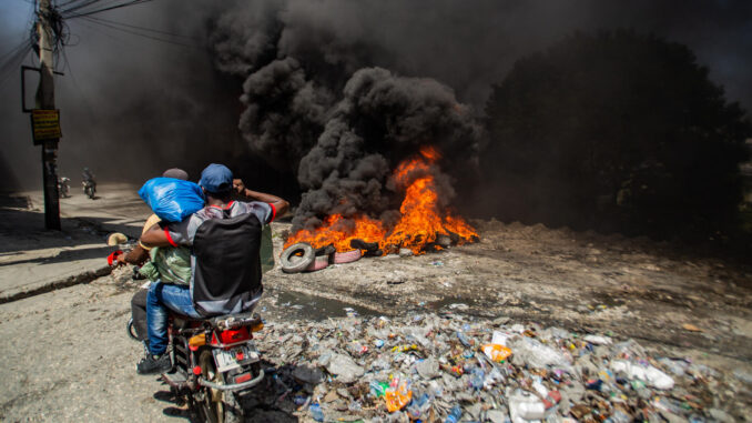 Dos hombres se movilizan en motocicleta frente a una barricada con fuego durante una protesta este lunes, en el barrio Solino, en Puerto Principe (Haití). EFE/ Mentor David Lorens
