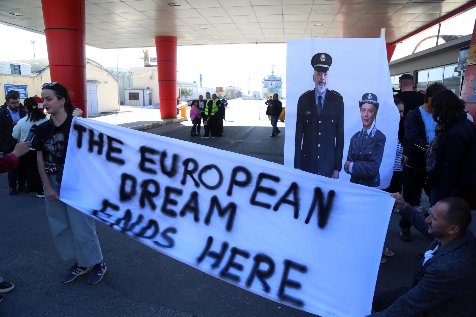 Activistas sostienen una pancarta con la primera ministra italiana, Giorgia Meloni, y el primer ministro albanés, Edi Rama, vestidos de policía, mientras el primer grupo de migrantes interceptados en aguas italianas llegan en un barco de la marina italiana a Shengjin, Albania, el 16 de octubre de 2024. EFE/EPA/MALTON DIBRA
