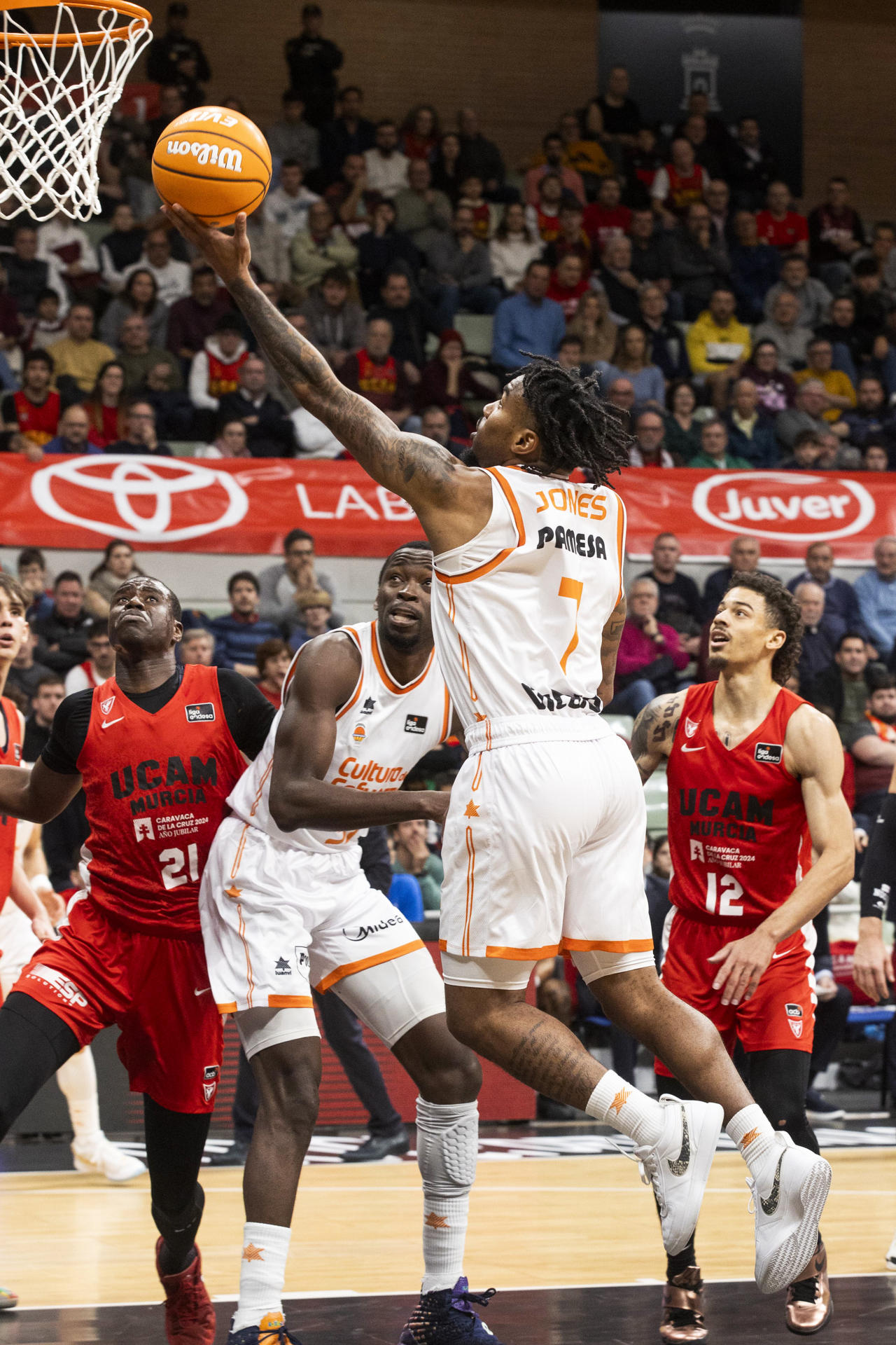 El base armenio de Valencia Basket Chris Jones (c) intenta encestar ante la mirada del pívot de UCAM Murcia Moussa Diagne (i), durante el partido de la décimo primera jornada de la liga Endesa jugado este domingo en el Palacio de los Deportes de Murcia. EFE/Marcial Guillén
