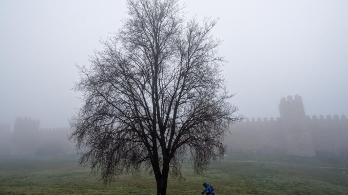 Una persona monta en bicicleta junto a la muralla de Ávila, entre la niebla, este martes. EFE/ Raúl Sanchidrián
