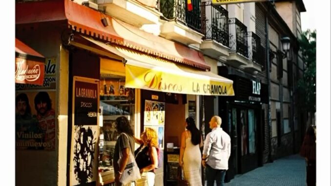 Vista urbana de Madrid mostrando espacios comerciales y callejeros con tiendas como Carrefour Express y La Gramola, capturada con efecto de grano cinematográfico