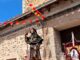 Estatua de San Roque decorada con flores y banderas frente a un edificio de piedra durante el desfile de las fiestas patronales de Valdecabras
