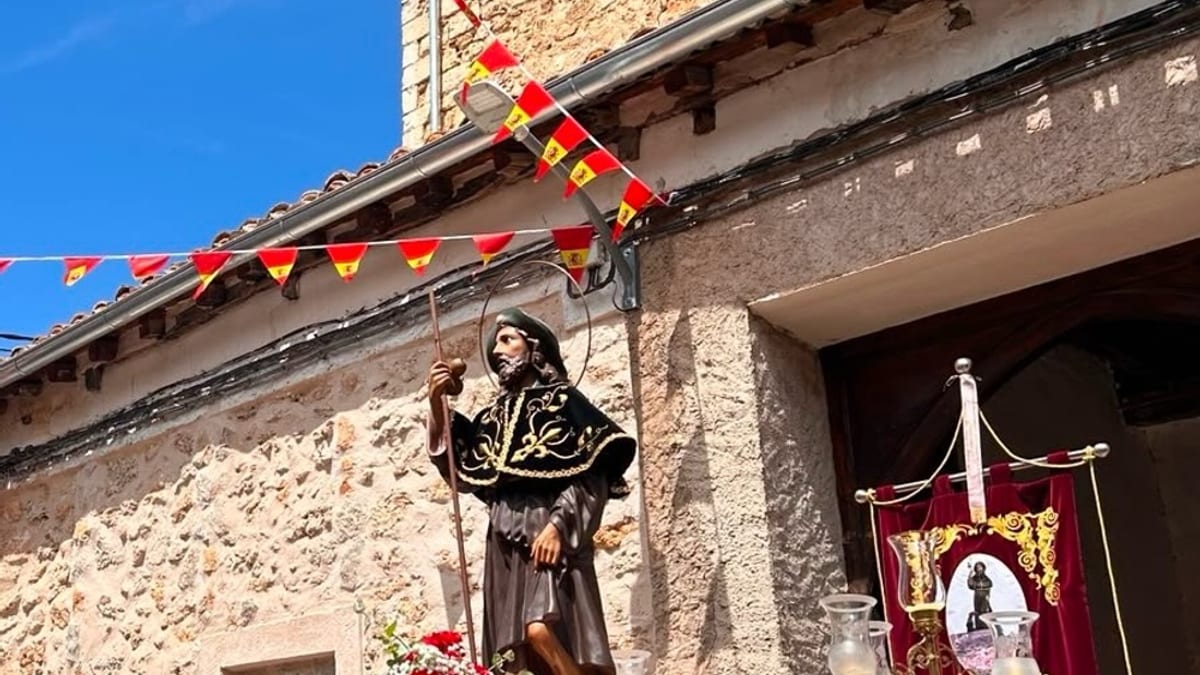 Estatua de San Roque decorada con flores y banderas frente a un edificio de piedra durante el desfile de las fiestas patronales de Valdecabras