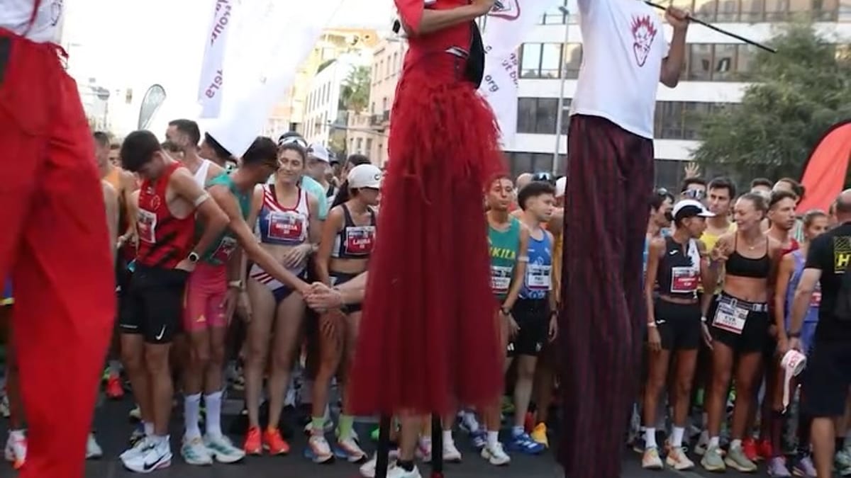Escena de la Cursa de la Mercè 2026 con corredores y performers en zancos en las calles de Barcelona
