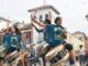 Participantes de la batucada Pinto Retumba tocando tambores en una plaza, vistiendo camisetas azules con logo amarillo