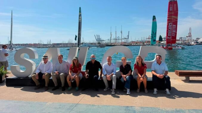 Grupo de personas en la playa frente a letras SU 100, con veleros y embarcaciones del equipo Emirates al fondo en Marina de València