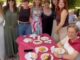 Reunión festiva al aire libre en la Asociación Parque Grande durante la celebración del Día de la Mujer en Alcorcón, con asistentes, mesa decorada con mantel rosa, comida y bebidas, e instalaciones decorativas con cintas de colores